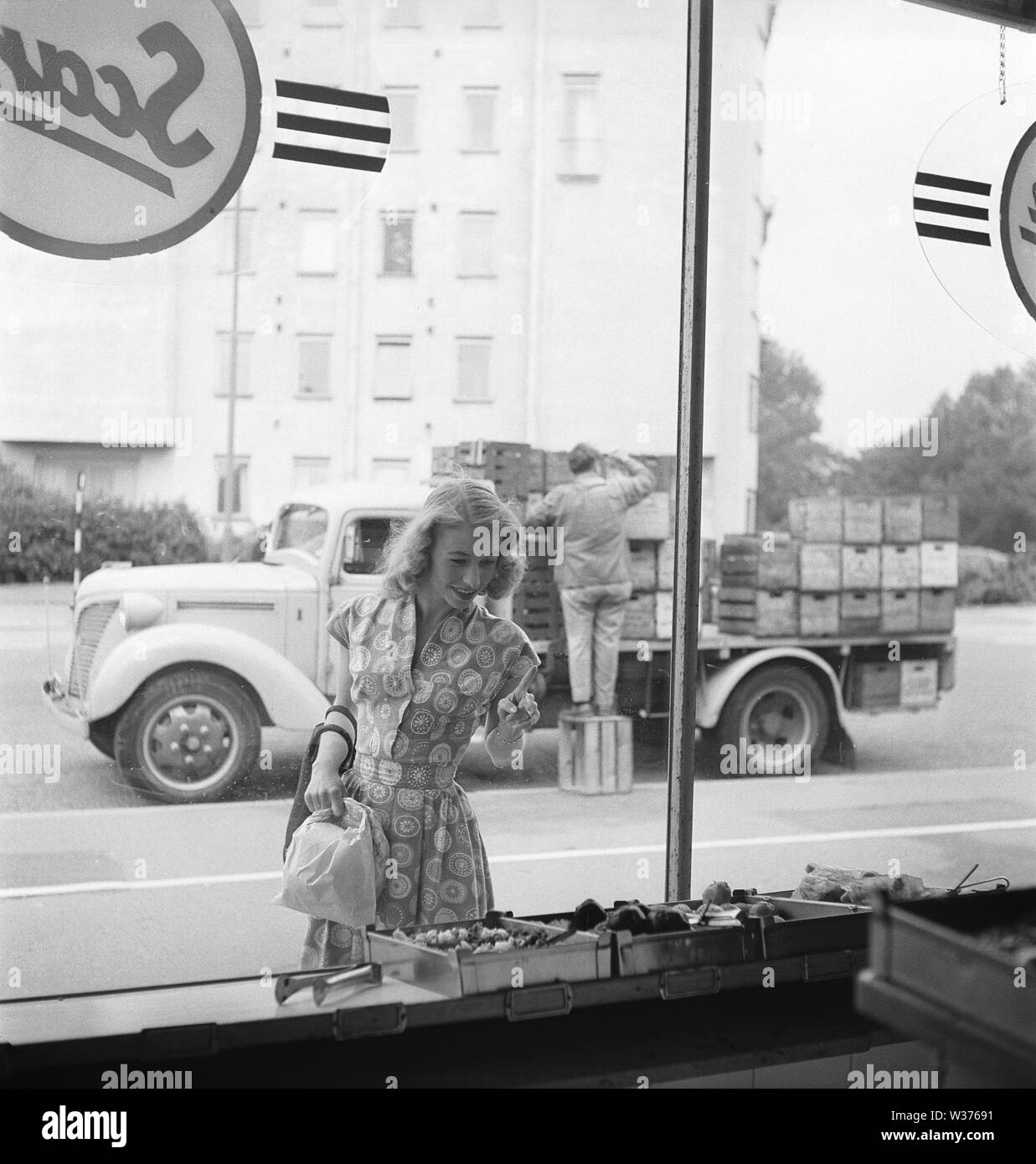 Window Shopping in den 1950er Jahren. Die junge Schauspielerin Erna Groth, 1931-1993, dargestellt außerhalb ein Lebensmittelgeschäft Fenster, in dem Sie zeigt mit dem Finger das Gemüse im Store Fenster zu wählen. Im Hintergrund wird ein Lkw mit Kästen Bier und andere Getränke für die Verteilung. Schweden 1952. Kristoffersson BG 68-3 Stockfoto