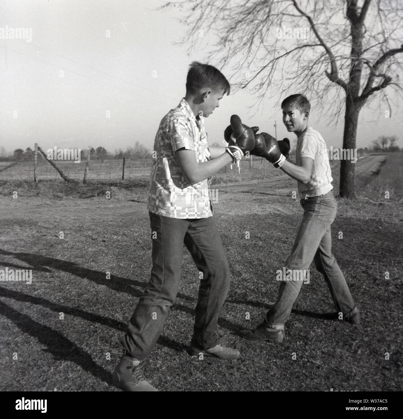 1960er Jahre, historisch, zwei ältere Teenager-Jungen, Brüder, vielleicht 16-18 Jahre alt, in Jeans und kurzärmeligen Hemden und mit Boxhandschuhen, die sich draußen auf einem Feld auf einer Farm, Mid-West, USA, gegenüberstehen. Stockfoto