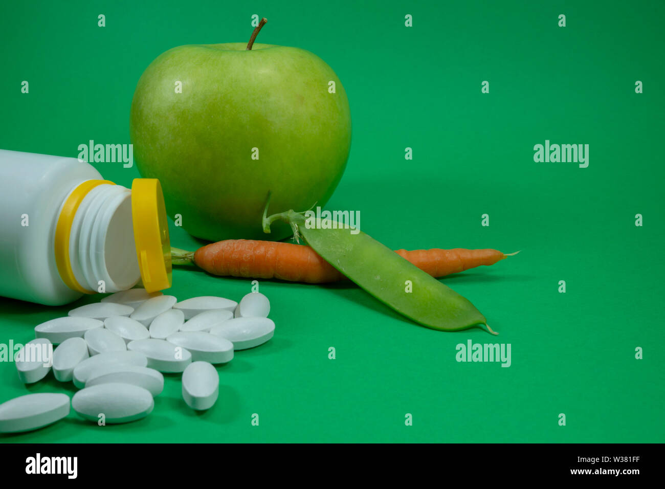 Pillen aus einer weißen Flasche in der Nähe von Apple, pea Pod und Karotte auf grünem Hintergrund. Healthcare, Diät oder Gewichtsverlust Konzept Stockfoto