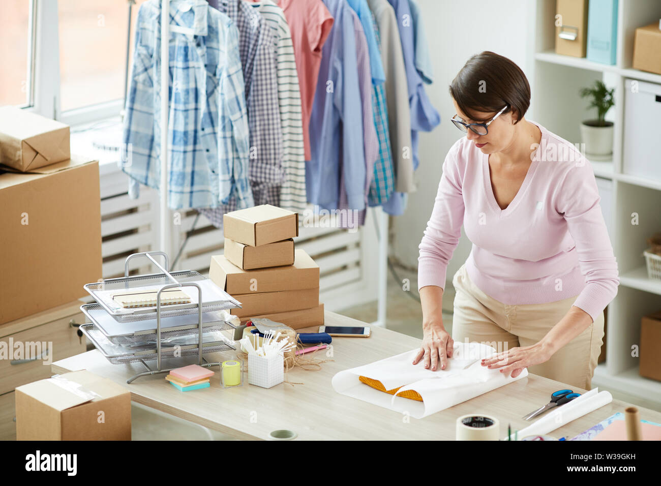 Junge brünette Frau Verpackung gefaltet Casualwear in Papier, während im Büro der Online Shop funktioniert Stockfoto