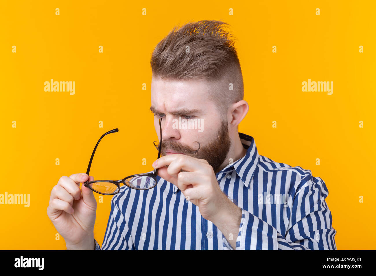 Junger hübscher Kerl ist mit Blick auf seine Brille während vor einem gelben Hintergrund. Das Konzept der Fehlsichtigkeit und Behandlung. Stockfoto