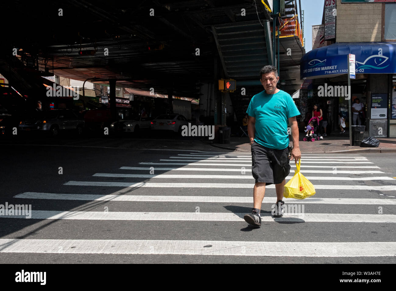 Ein Mann mittleren Alters mit einer Einkaufstasche Kreuze 96th Street unter der erhöhten #7 Zug auf Roosevelt Ave in der Corona, Queens, New York City. Stockfoto