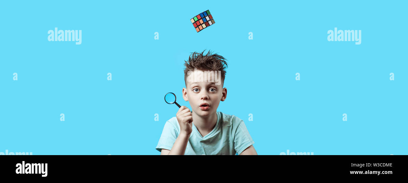 Ein Junge in einem hellen t-shirt hält eine kleine Lupe in der Hand und einen Rubik's Cube fällt von oben. Stockfoto