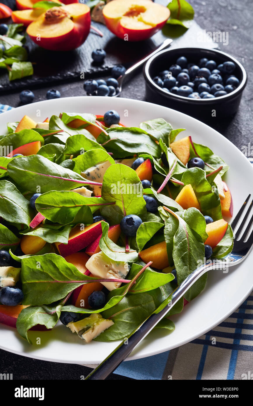 Nahaufnahme der Salat mit frischen Mangold Blätter, Pfirsich, Blaubeeren, Stücke von blauen Schimmel Käse serviert auf einem weißen Teller auf einen konkreten Tisch mit Zutaten Stockfoto