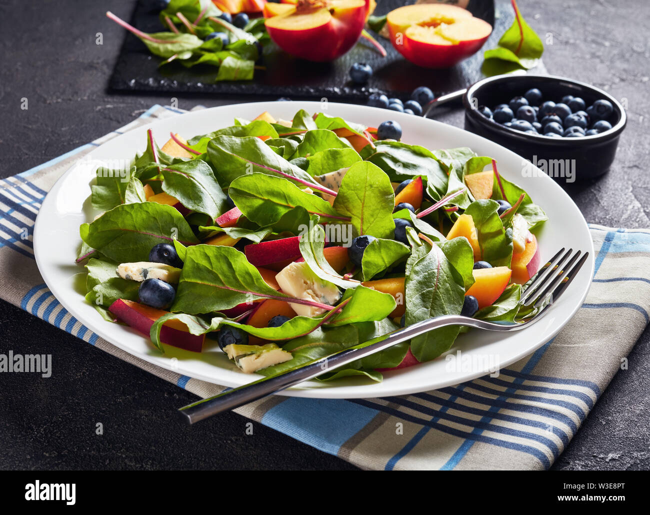 Salat mit frischen Mangold Blätter, Pfirsich, Blaubeeren, Stücke von blauen Schimmel Käse serviert auf einem weißen Teller auf einen konkreten Tisch mit Zutaten, horizontal Stockfoto