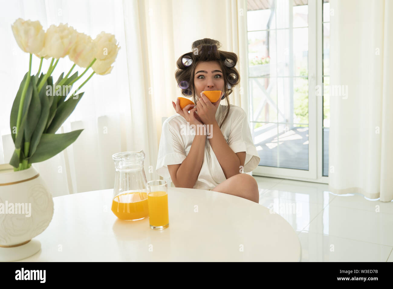 Schöne junge brünette Frau in Lockenstäbe beißen orange und orange in einem anderen Hand in helle Küche Stockfoto