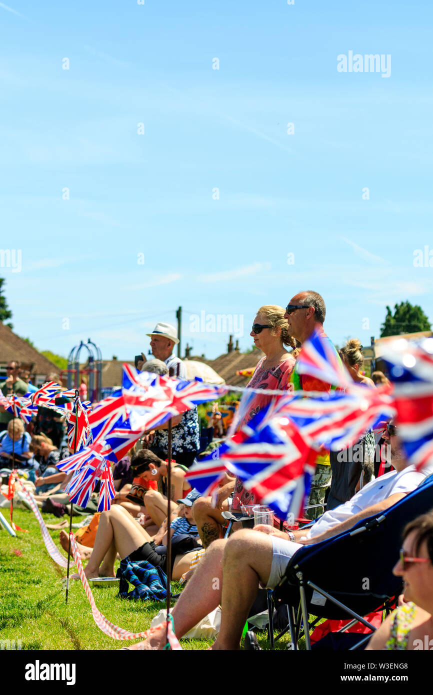 Union Jack bunting flattern im Wind, bei der die Zeile der Menschen dahinter stehen und sitzen gerade unseen Event. Strahlender Sonnenschein. Negativer Platz. Stockfoto