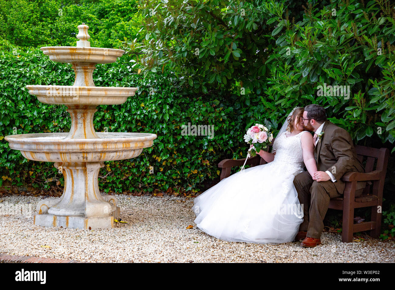Braut und Bräutigam küssen auf einer Bank neben einem alten Brunnen Stockfoto