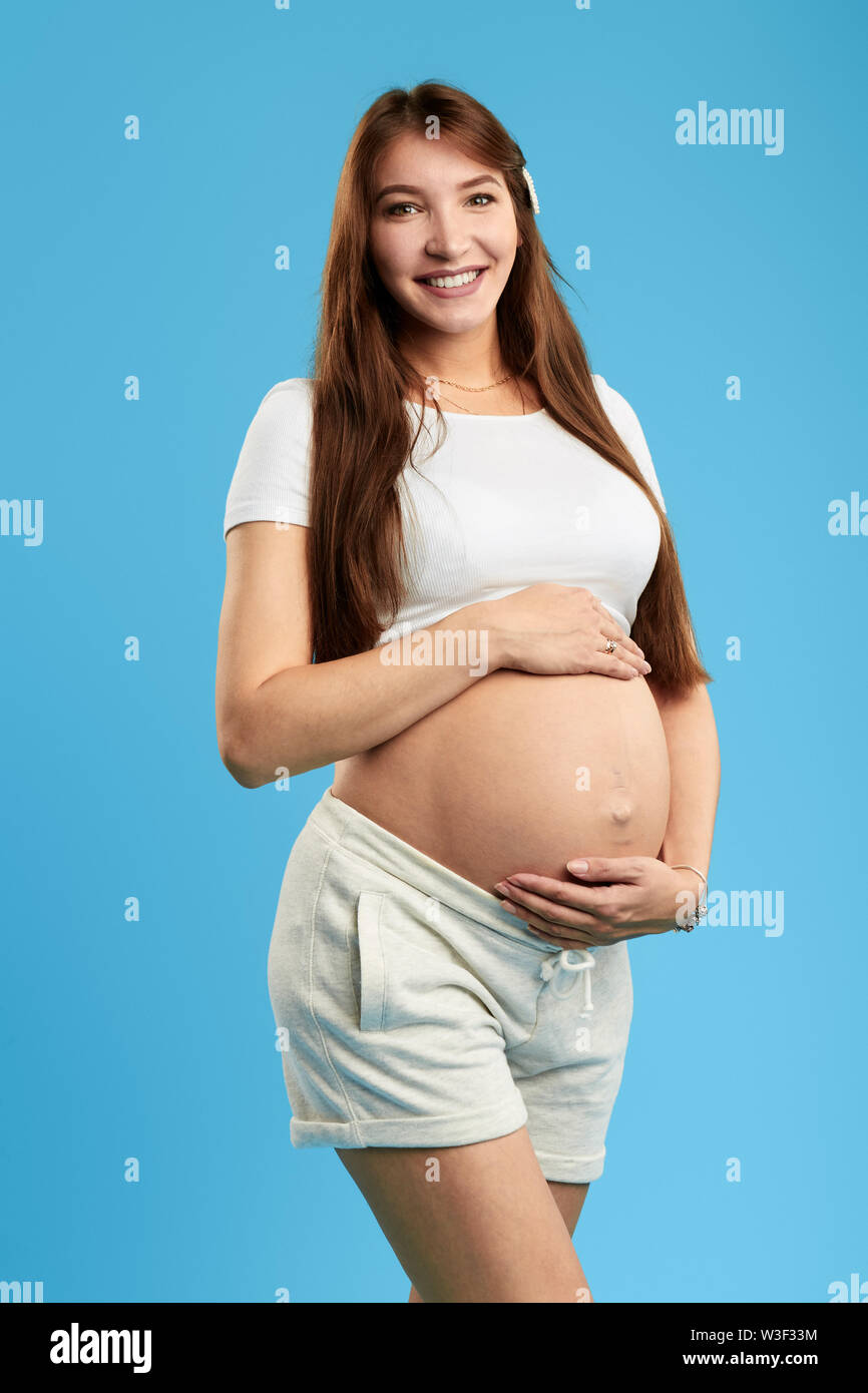 Positive schöne junge Frau mit großen Bauch in die Kamera schaut. Glück, neues Leben, isoliert blauen Hintergrund. studio Shot, erben und Nachfolger Stockfoto