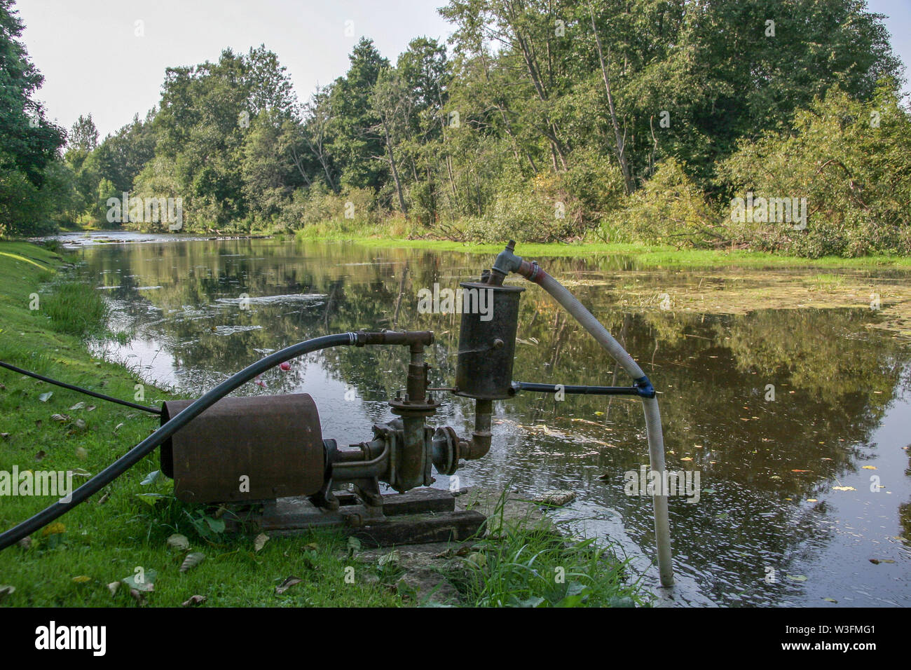 Eine große alte Wasserpumpe steht am Ufer des Flusses und pumpen Wasser.  Der Fluss ist mit Gras bewachsen. Bäume wachsen auf dem Ufer  Stockfotografie - Alamy