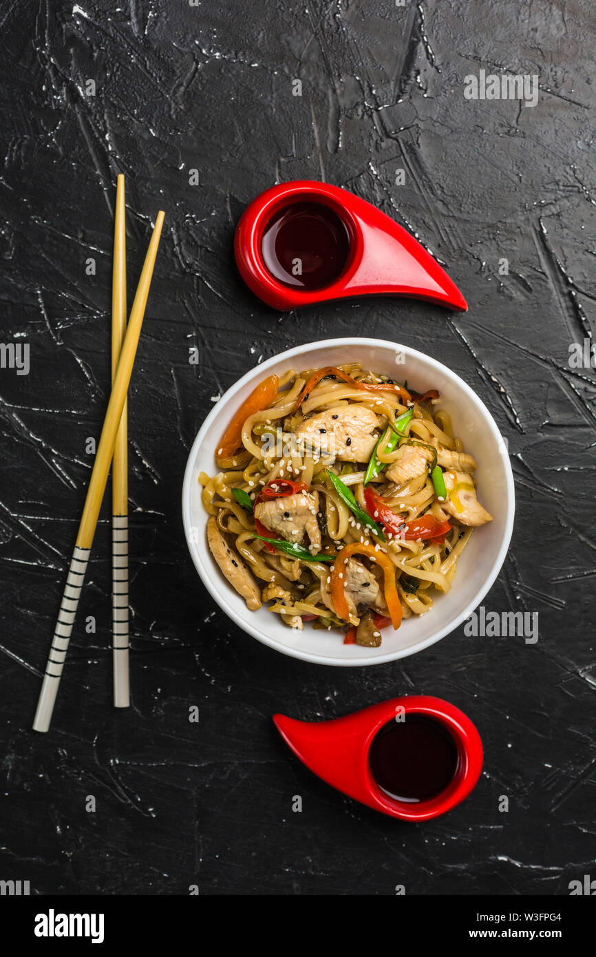 Chinesische Nudeln in eine Schale mit Hähnchen und Gemüse mit Stöcken. Wok Essen aus dem Restaurant auf einem dunklen Hintergrund von oben gesehen. Stockfoto