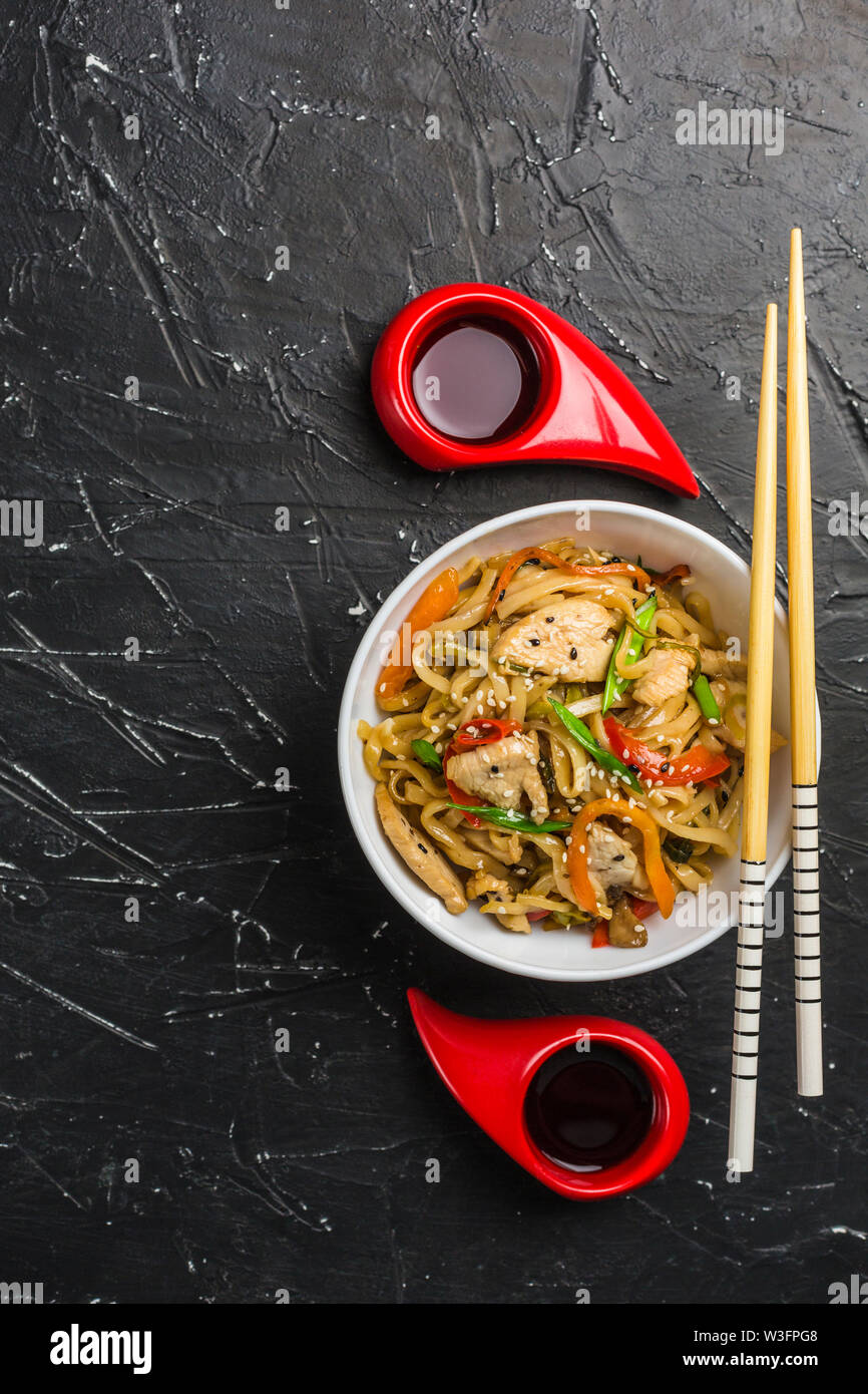 Chinesische Nudeln in eine Schale mit Hähnchen und Gemüse mit Stöcken. Wok Essen aus dem Restaurant auf einem dunklen Hintergrund von oben gesehen. Stockfoto