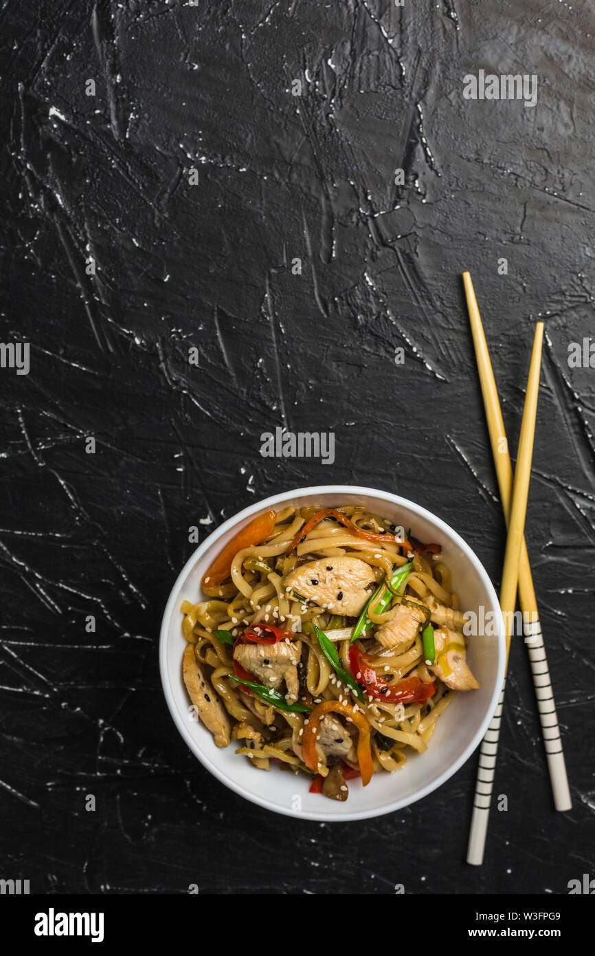 Chinesische Nudeln in eine Schale mit Hähnchen und Gemüse mit Stöcken. Wok Essen aus dem Restaurant auf einem dunklen Hintergrund von oben gesehen. Stockfoto