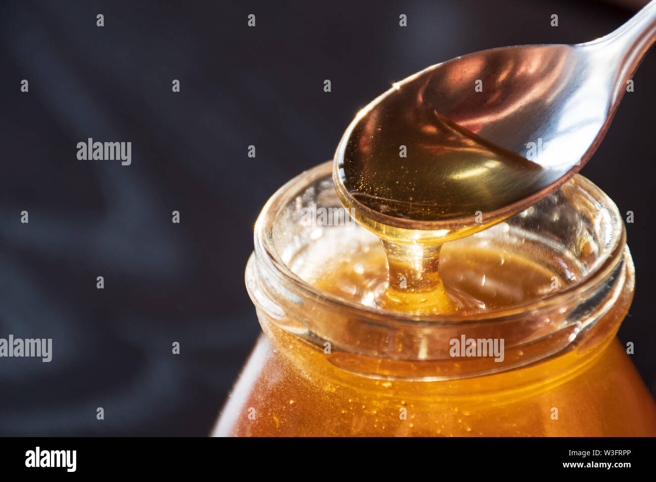 Delicious Organic Natural Bio Honig gießt der Löffel in ein Glas Glas auf Holztisch mit Reflexion der Sonne, schwarzer Hintergrund, Nahaufnahme Stockfoto