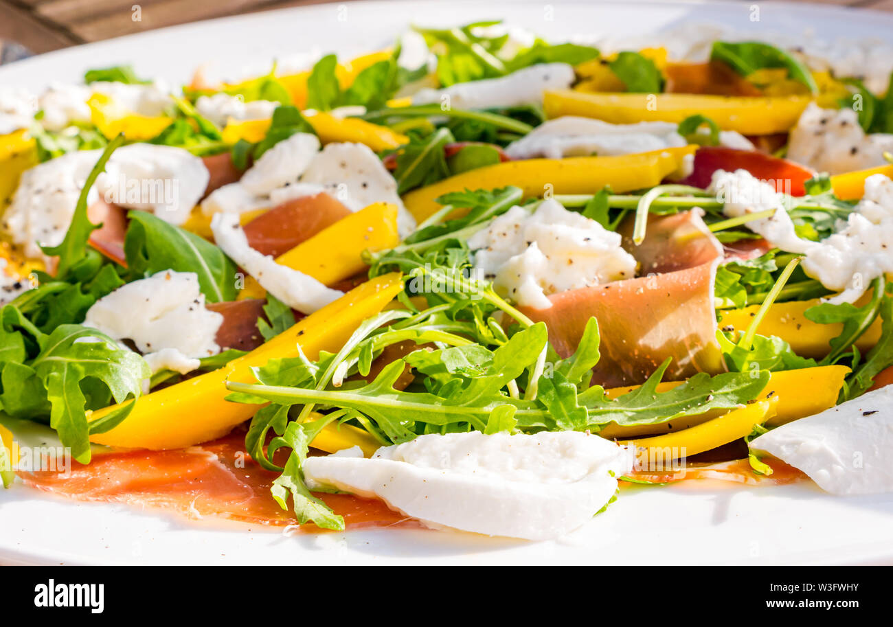 Sommer Essen: Salat essen im Freien mit Mango Slices, Rakete, Mozzarella und Parmaschinken auf einer weißen Platte Stockfoto