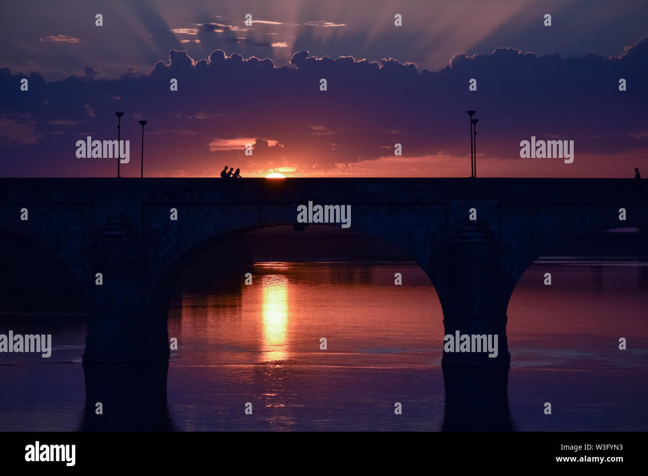 Einen atemberaubenden Sonnenuntergang mit Licht, das durch die Wolken und einem bunten Himmel. Im Vordergrund eine Brücke, einer Stadt in der Hintergrundbeleuchtung. Stockfoto