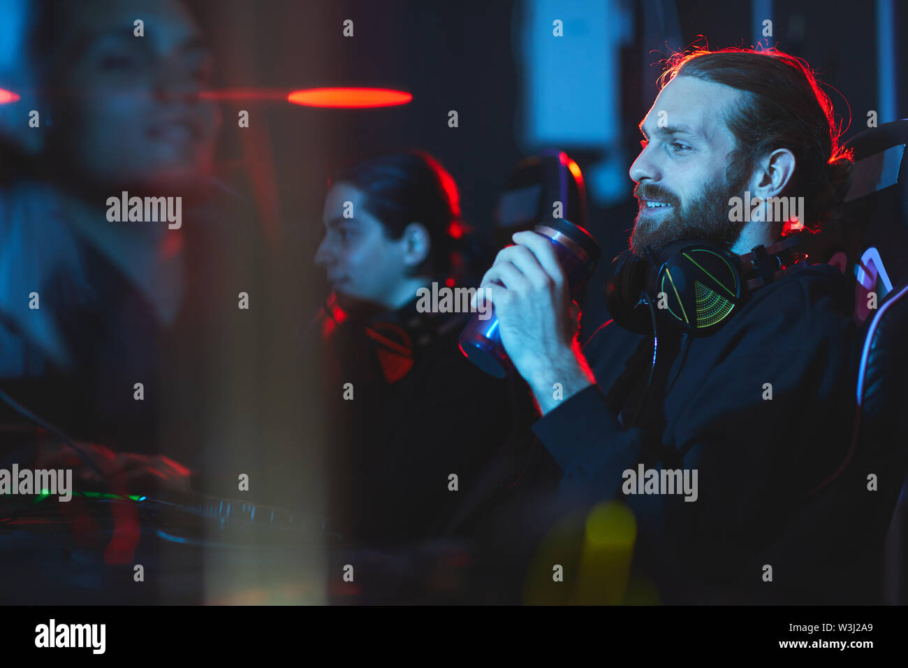 Zufrieden lächelnd junge Bartgeier gamer mit Kopfhörer auf dem Hals sitzen in bequemen Sessel und trinken Energie Getränke beim Spielen video game Stockfoto