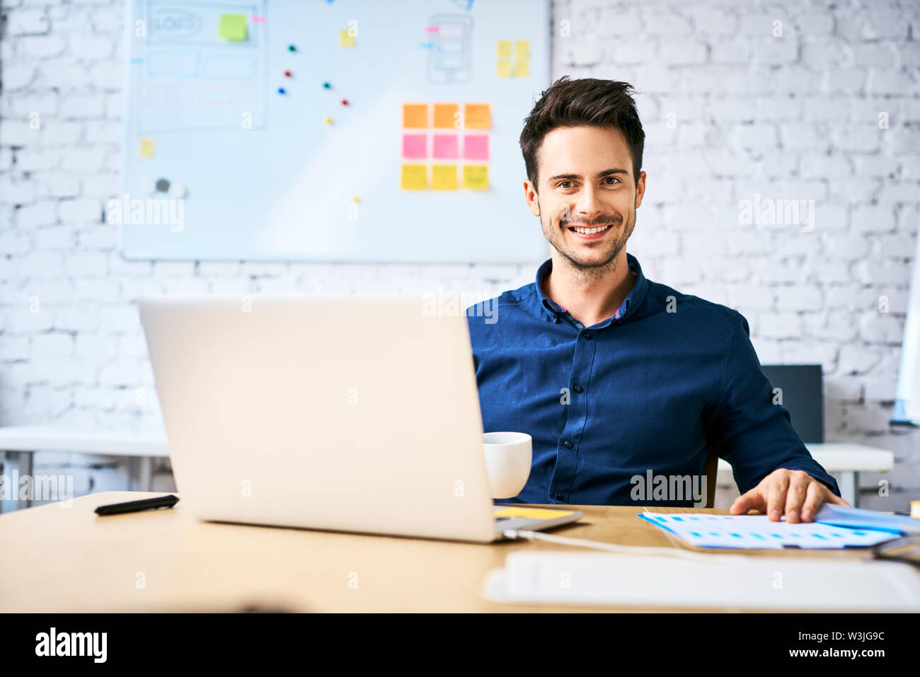Startup Unternehmer am Schreibtisch mit Blick auf die Kamera sitzen beim Kaffee trinken und Arbeiten mit Laptop und Notizen Stockfoto