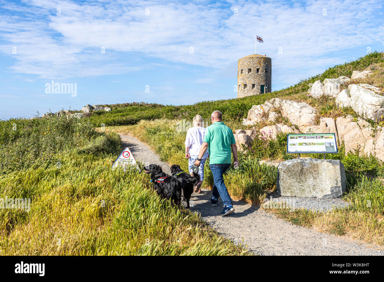 Ein Paar mittleren Alters, ihre Hunde auf dem Küstenweg neben Schlupfloch Turm Nr. 5 L'Ancresse (Nid de l'Herbe, Vale), Guernsey, Kanalinseln, Großbritannien Stockfoto