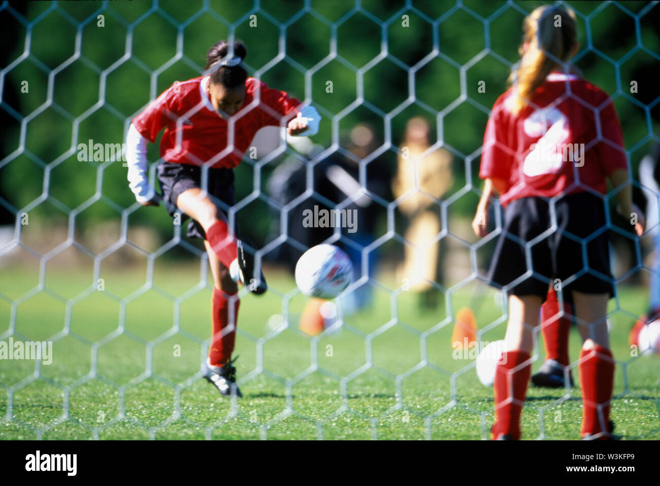 Ein Mädchen tritt ein Fußball während der Torwart steht warten, wie von hinter dem Tor gesehen. Stockfoto