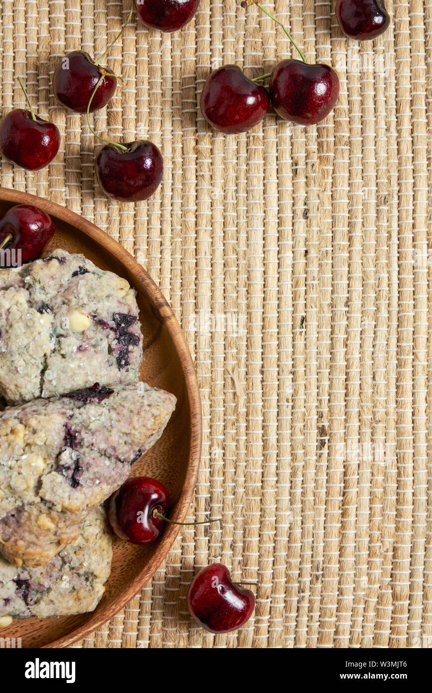 Holzplatte mit Cherry und weiße Schokolade Gebäck auf einem Gewebe Tischset Hintergrund mit frischen Kirschen vertikale Stockfoto