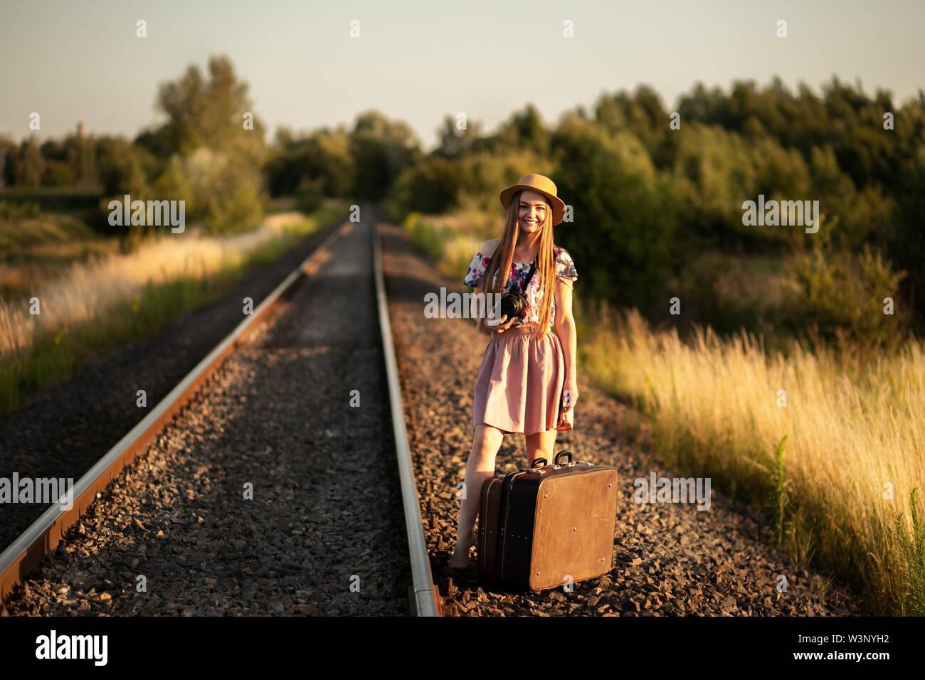 Schlanke Mädchen in einem Strohhut mit Koffern auf den Eisenbahnschienen, mit einer Kamera, lächelnd und Träumen. Das Konzept der Reisen, Glück, freedo Stockfoto