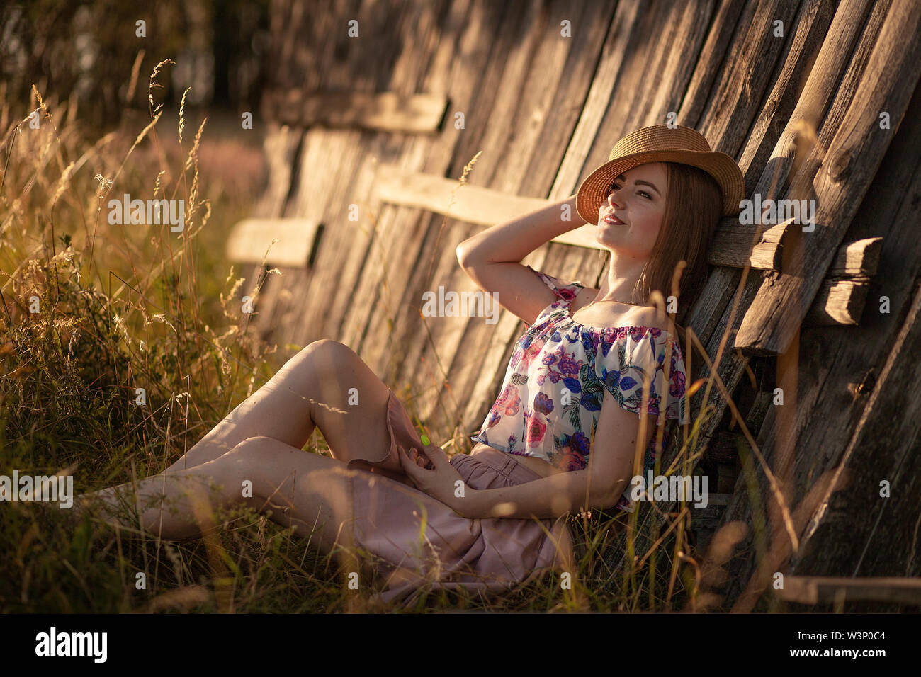 Schönen schlanken Mädchen mit langem Haar in einem Strohhut sitzen in der Nähe von einem Holzzaun und Träumen. Sommer am Abend bei Sonnenuntergang. Liebe Konzept Glück, Jugend Stockfoto