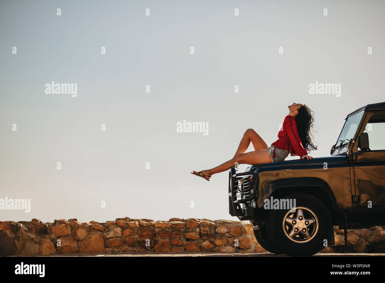 Seitenansicht des junge Frau sitzt auf dem Auto Motorhaube und oben zu schauen. Weibliche eine Pause auf der road trip. Stockfoto
