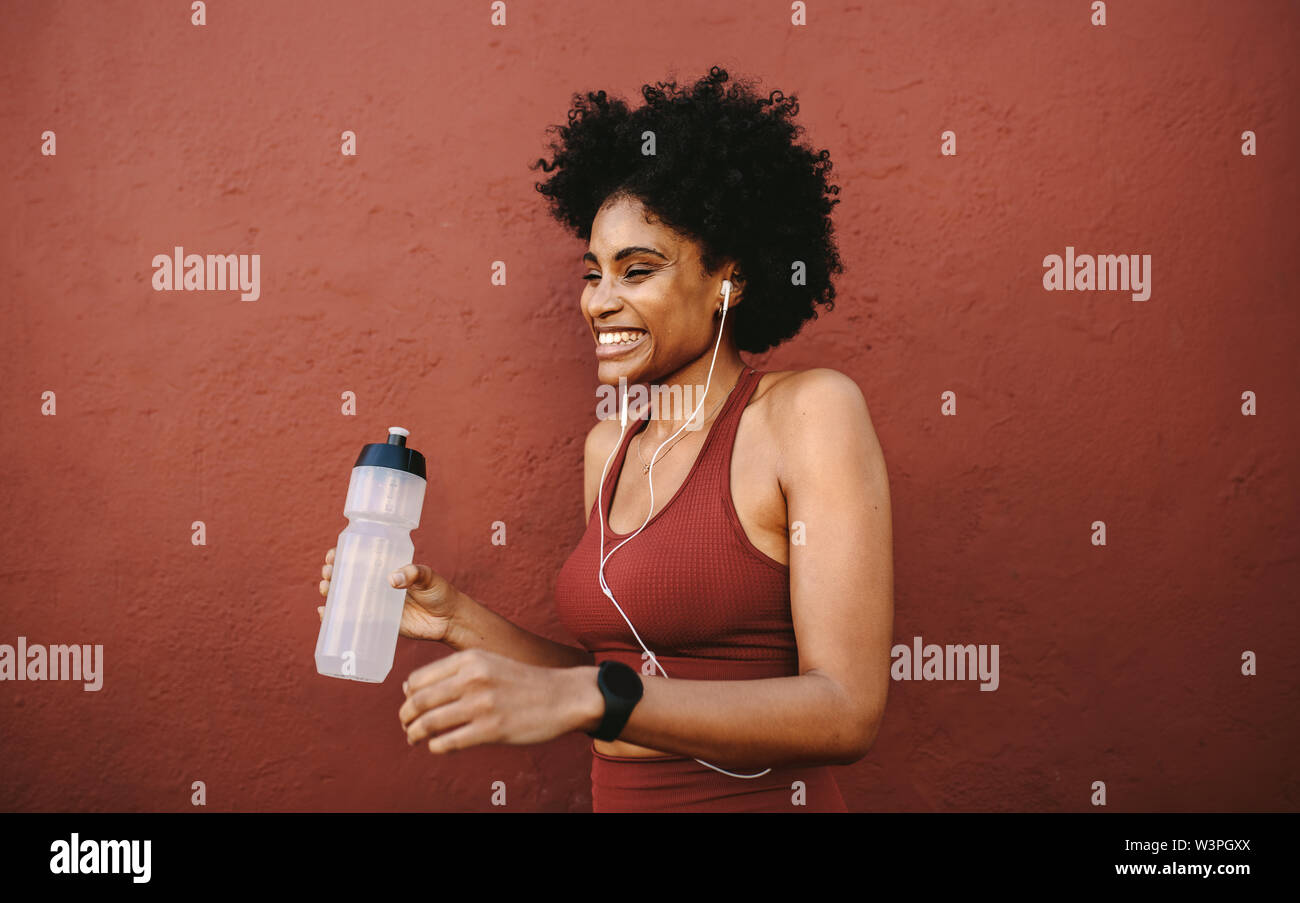 Fitness-Frau steht mit Wasserflasche und lächelt. Läuferin, die sich nach dem Workout ausruht, sich an eine Wand lehnt und wegblickt und lächelt. Stockfoto