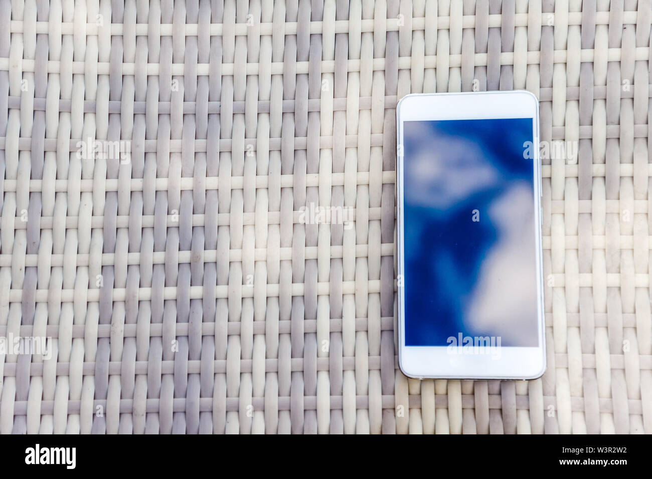 Reflexion der blauen Himmel mit Wolken im Telefon - Smartphone, das auf einem hellen Rattanmöbeln Tabelle vor dem Hintergrund liegt. Blur und Abstraktion Stockfoto