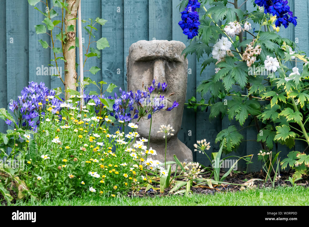 Ein betonwerkstein Garten Ornament in Form von einem der berühmten Osterinsel Köpfe sitzt zwischen Blumen in einem privaten Garten gelegen Stockfoto