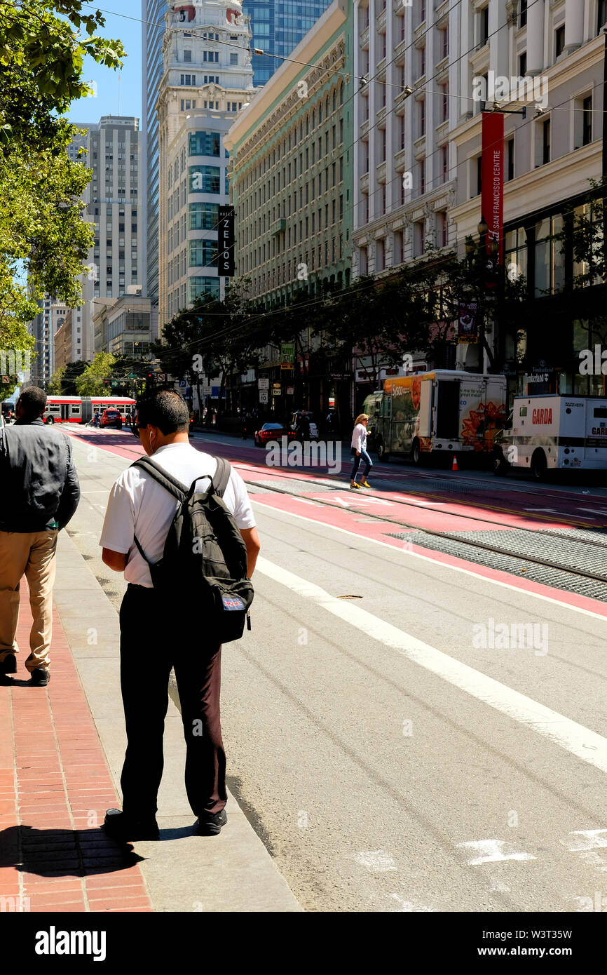 Hispanic Mann mit einem Rucksack auf den Bus warten auf der Market Street in der Innenstadt von San Francisco, Kalifornien auf einer sonnigen und ruhigen Sommertag. Stockfoto
