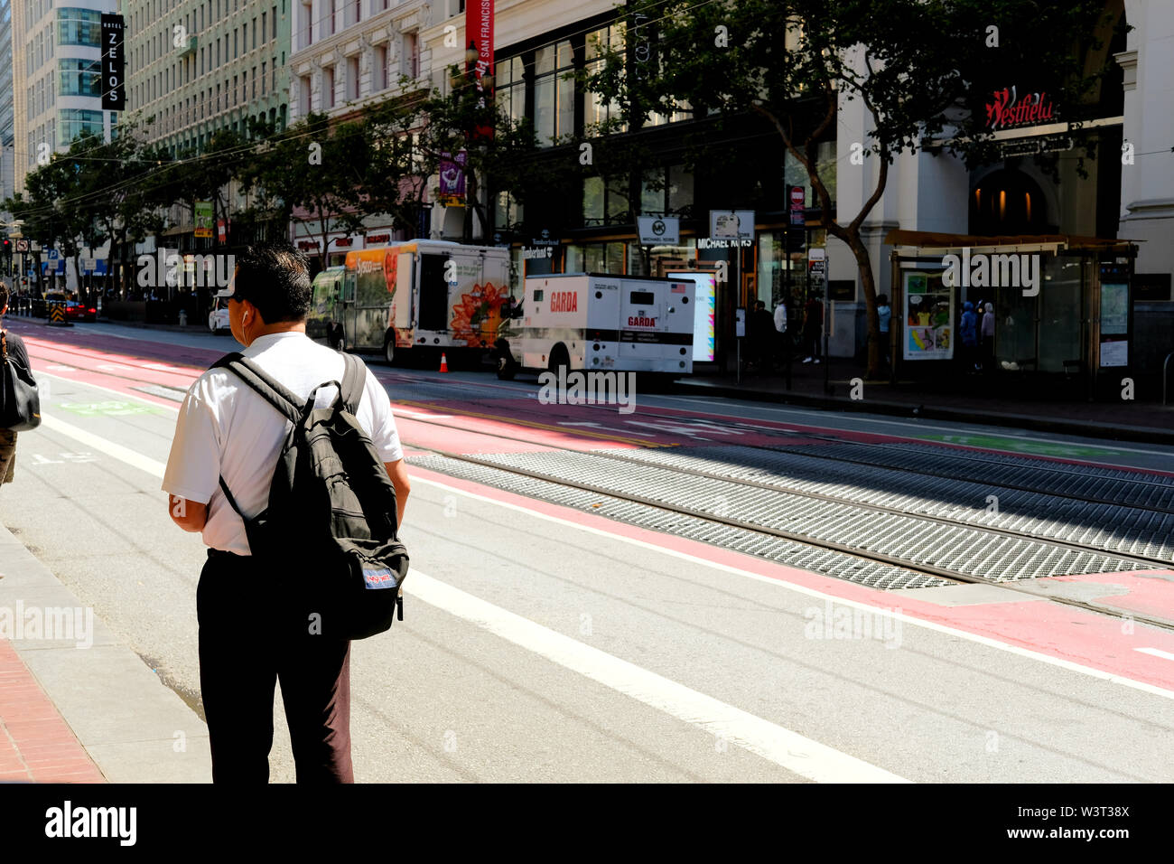 Hispanic Mann mit einem Rucksack auf den Bus warten auf der Market Street in der Innenstadt von San Francisco, Kalifornien auf einer sonnigen und ruhigen Sommertag. Stockfoto