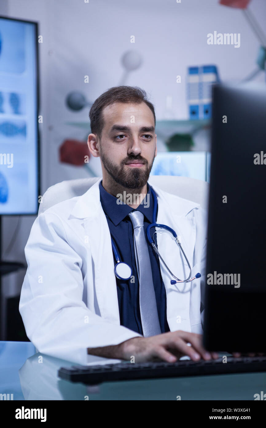 Hübscher junger Bartgeier Arzt sie in seinem Computer im Krankenhaus. Arzt im weißen Mantel mit einem Stethoskop. Stockfoto