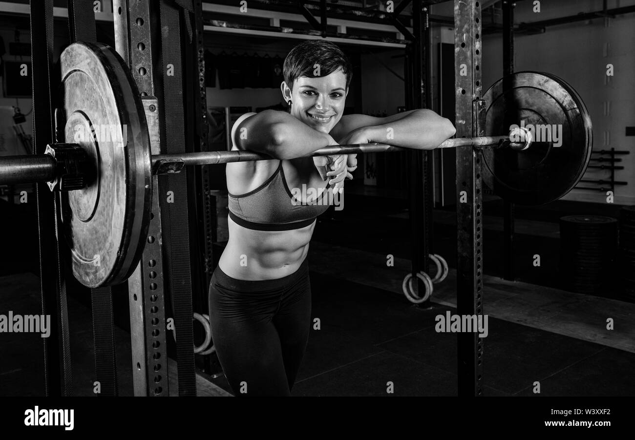 Schwarz-weiß-Porträt eines attraktiven weiblichen Athleten mit starken abs. Die athletische und lächelnd kurzhaarige Frau lehnt sich an die Hanteln. Stockfoto