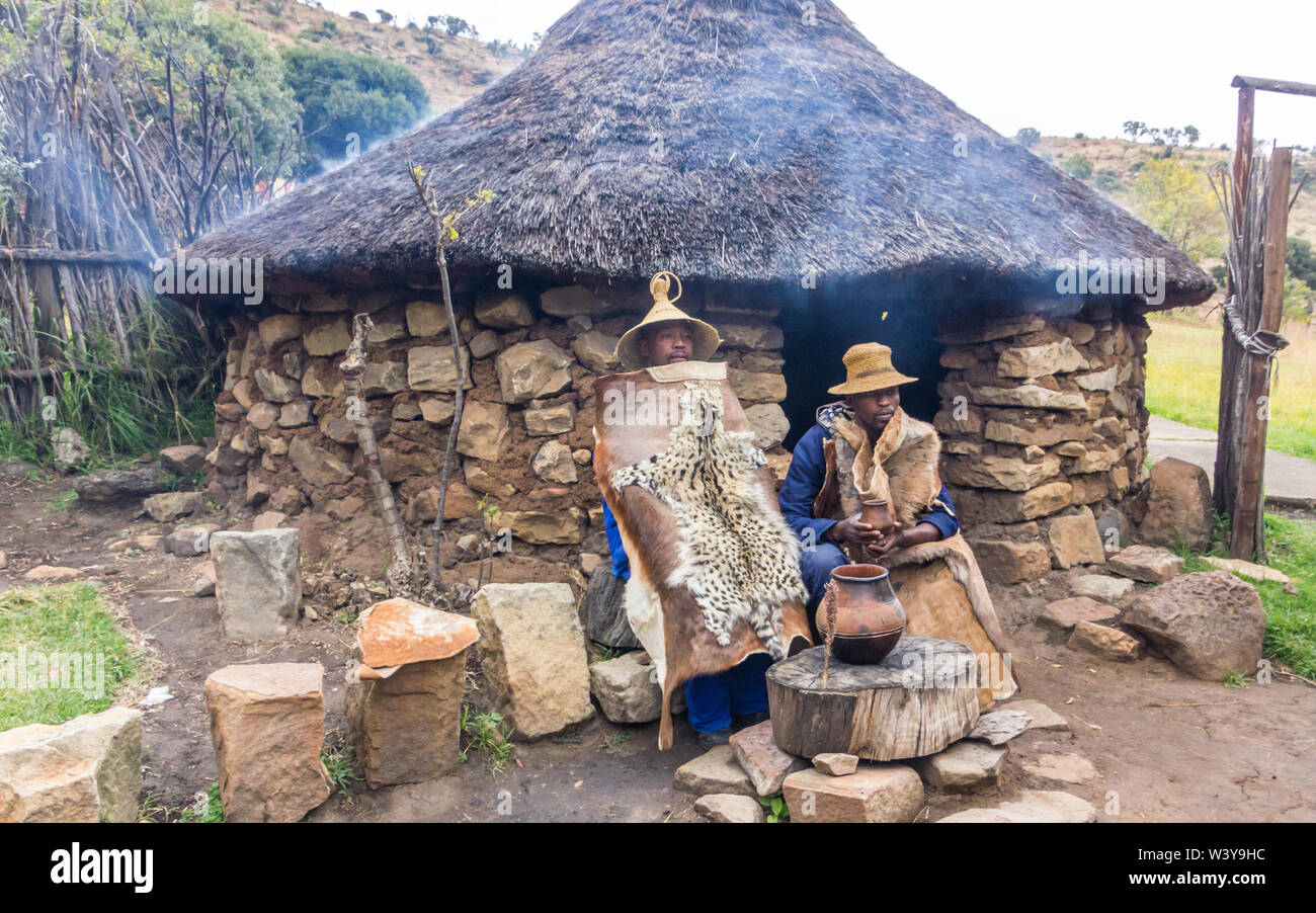 Basotho Cultural Village ländliche Szene mit zwei schwarze afrikanische Männer in traditionellen Felle oder Häute außerhalb einer Stein Thatch rondawel oder Hut gekleidet Stockfoto
