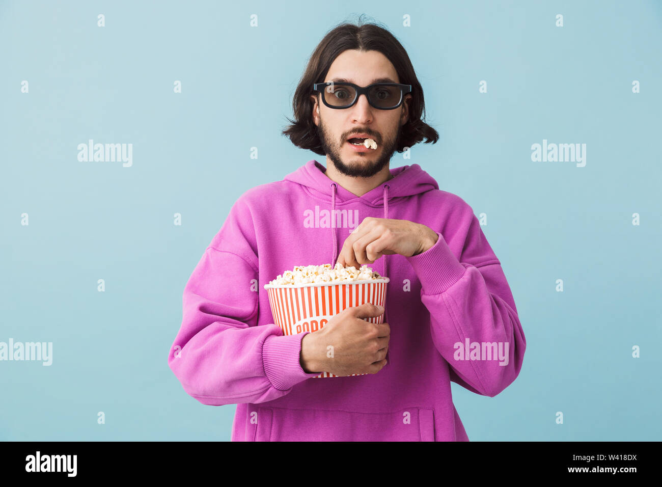 Portrait einer aufgeregt junge Bartgeier brünett Mann hoodie stehend über blauen Hintergrund isoliert, essen Popcorn, watching Film in 3D-Brille Stockfoto