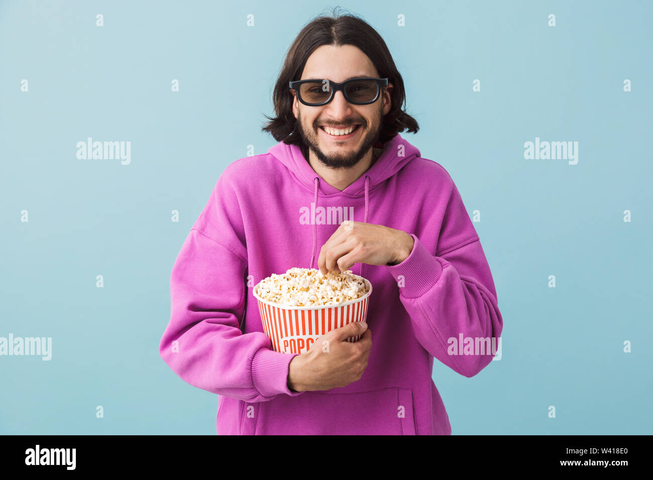 Portrait einer aufgeregt junge Bartgeier brünett Mann hoodie stehend über blauen Hintergrund isoliert, essen Popcorn, watching Film in 3D-Brille Stockfoto