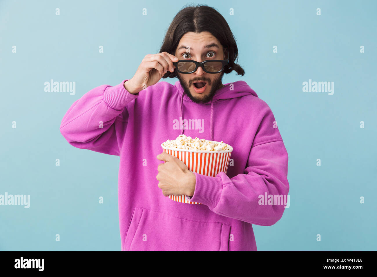 Portrait einer aufgeregt junge Bartgeier brünett Mann hoodie stehend über blauen Hintergrund isoliert, essen Popcorn, watching Film in 3D-Brille Stockfoto