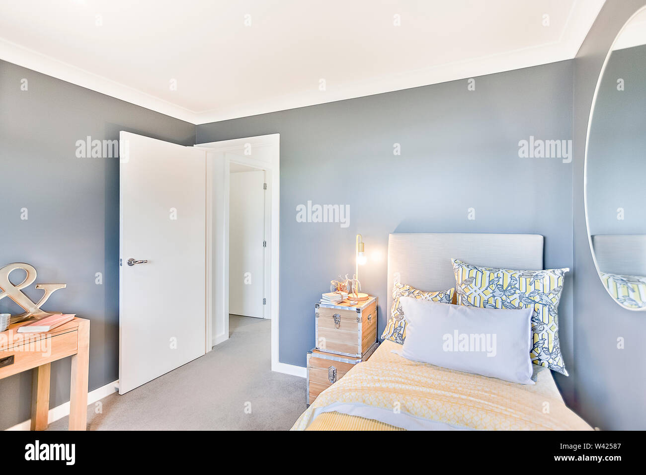 Modernes Schlafzimmer mit offener Tür und Wand Spiegel auf den hellblauen Wand gibt es Kissen auf den gelben Decke neben Holzkiste Lampen auf dem Teppich f Stockfoto
