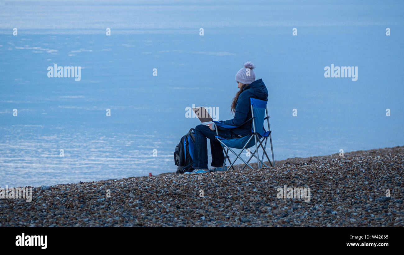 Eine junge Frau sitzt auf einem Stuhl an einem peeble beach Remotes Arbeiten mit Ihrem Laptop Stockfoto