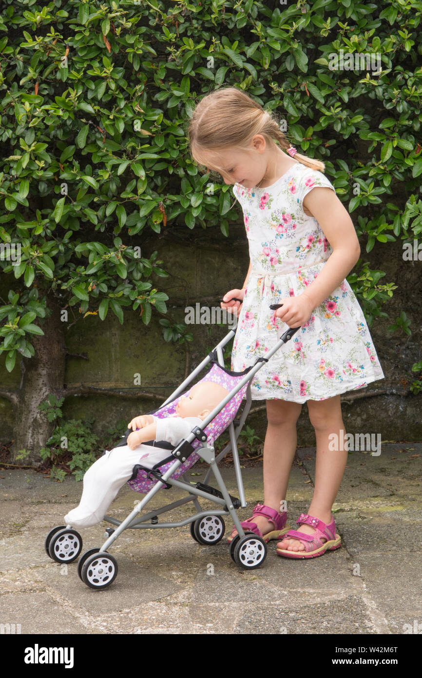 Drei Jahre alten Kind, junge Mädchen in der hübschen Kleid, blonde Haare in Pigtails spielen mit Puppe in Spielzeug Kinderwagen, draußen im Garten, Großbritannien Stockfoto