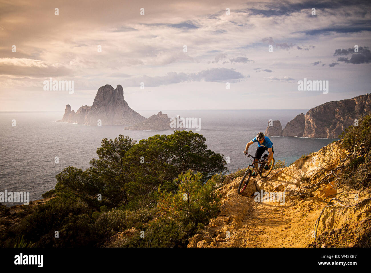 Ein Mann reitet ein Mountainbike entlang einer felsigen Trail an der Westküste von Ibiza. Es Vedrá Insel und das Mittelmeer sind im Hintergrund. Stockfoto