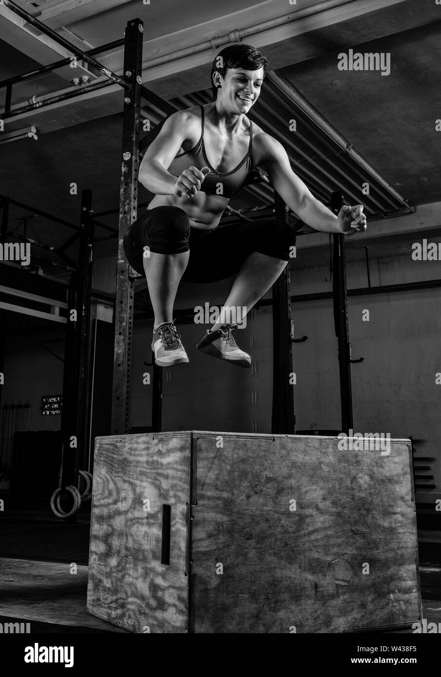 Eine athletische kurze behaarte Frau mit starken abs tut, springt in ein Fitnessstudio. Die weiblichen Athleten lächelt und springen auf die Box. Schwarz und Weiß. Stockfoto