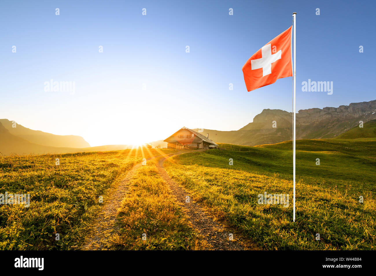 Kleine Straße zu einem Schweizer Chalet oder Bauernhof in der Berglandschaft bei Sonnenaufgang mit Sun Star führenden und eine wehende schweizer Fahne im Vordergrund. Stockfoto