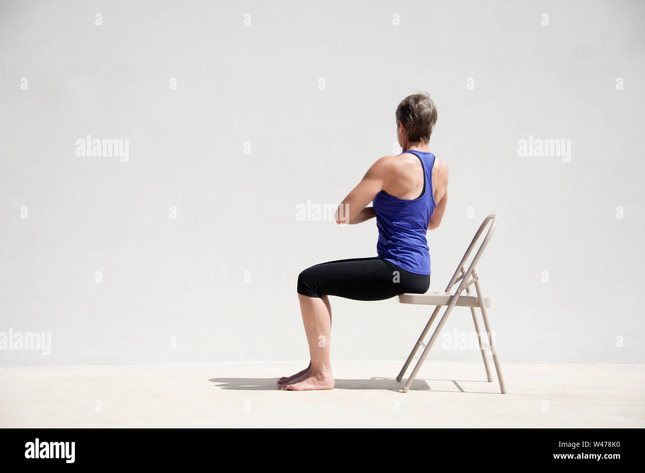 Reifer senior Frau practing Sanfte restorative Yoga in einem weißen Hintergrund darstellt. Sie hat mit erhobenen Händen, während er auf eine spezielle Therapie Stuhl. Stockfoto