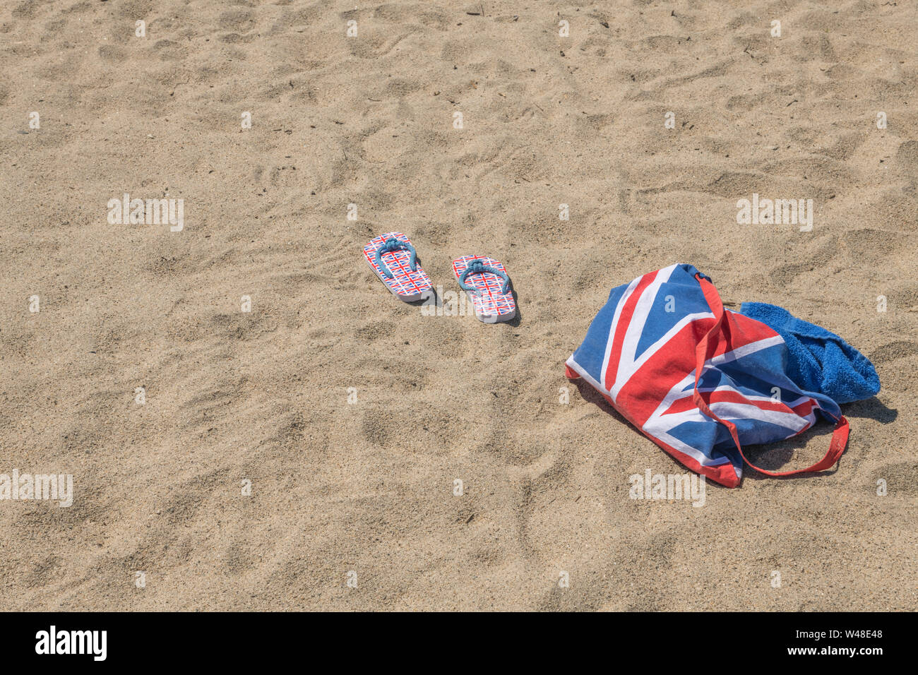 Union Jack Strandtasche & Flip-Flops am Sandstrand - für 2021 Aufenthalte in Großbritannien, Urlaub zu Hause, Aufenthalt in Cornwall, Badeurlaub, Flip-Flop-Schuhe. Stockfoto