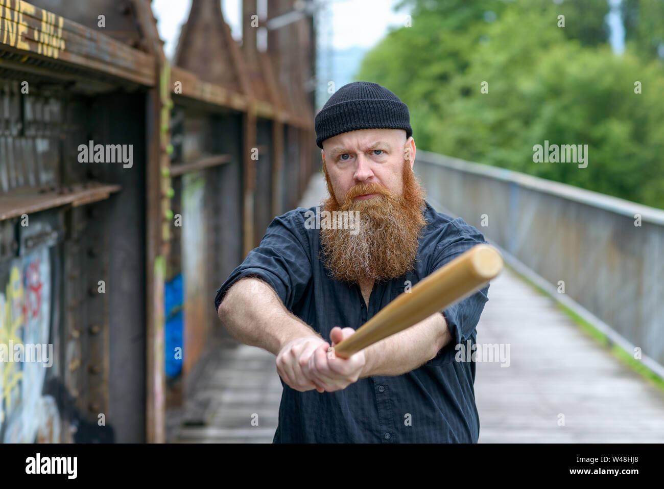 Aggressive bärtiger Mann auf gestrickte Mütze Hut schwingen einen Baseballschläger in die Kamera mit einem wütenden Ausdruck im Freien auf einer Brücke Stockfoto