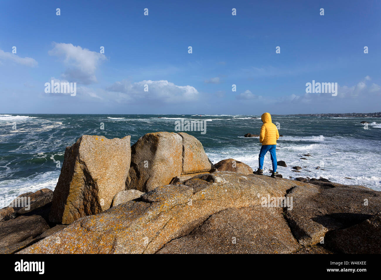 Junge in gelber Jacke stehend auf Granit Felsen beobachten große Wellen aus dem Atlantik auf schroffe Küstenlandschaft in der Bretagne, Frankreich Stockfoto