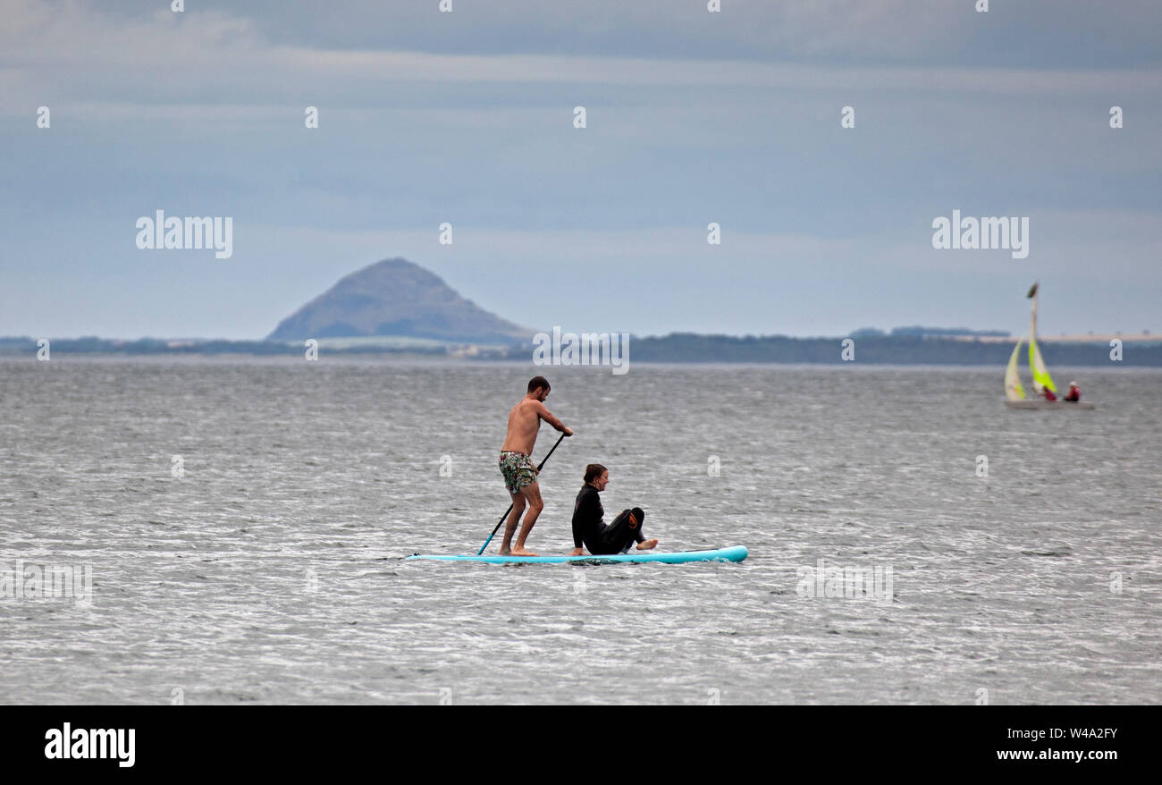 Portobello, Edinburgh, Schottland, Großbritannien. Juli 2019 21. Sportliche Aktivitäten gerade weg von Portobello Beach ein Mann Stand up Paddle Boarding mit weiblichen Passagier mit Berwick Gesetz und in einem Beiboot hinter sich. Stockfoto
